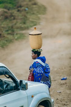 A Kenyan woman carrying a basket on her head, walking beside a vehicle on a dirt road.